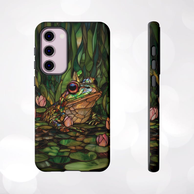 May include: A phone case with a stained glass design featuring a green frog with red eyes and pink flowers. The case is black and has a cutout for the camera lens.