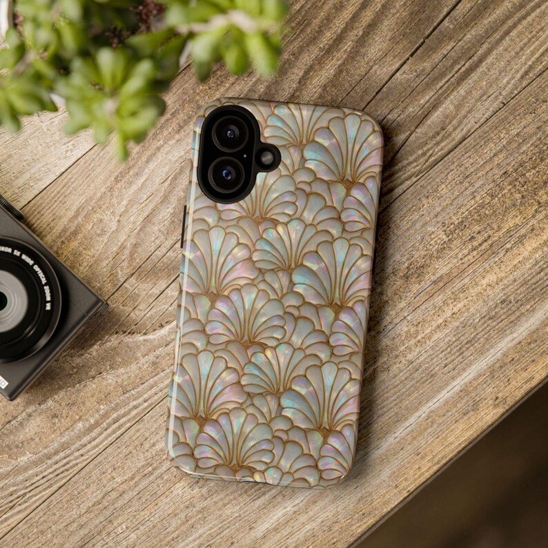 May include: A smartphone case with a pearlescent, fan-shaped pattern in shades of white, gold, and iridescent hues. The case is lying on a wooden surface, showcasing a decorative design. The phone case is a protective accessory.