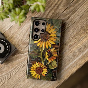 May include: A phone case with a stained glass design featuring sunflowers in shades of yellow, orange, and brown. The case has a cutout for the phone's camera lens.
