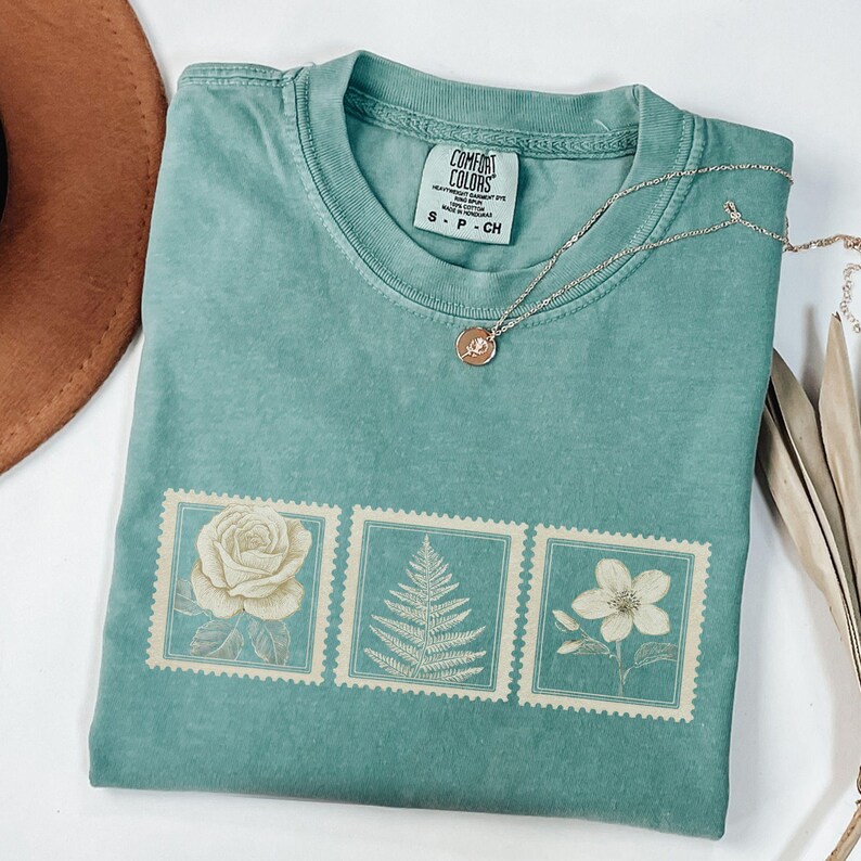 May include: A teal Comfort Colors t-shirt featuring three postage stamp designs with botanical illustrations of a rose, fern, and flower. A silver necklace with a pendant rests on the shirt. The shirt is folded and has a small tag.