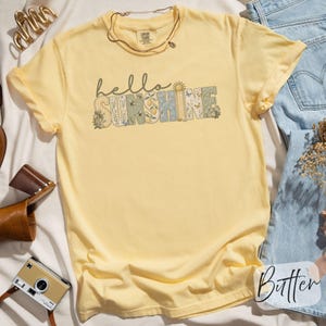 May include: Yellow t-shirt with the words "hello SUNSHINE" in a decorative font. The word "SUNSHINE" has a floral and patterned design. A gold necklace is draped over the shirt. The shirt is folded on a white surface.
