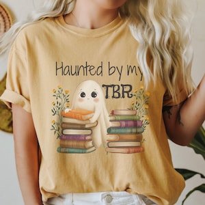 May include: Mustard yellow t-shirt with the phrase "Haunted by my TBR" and a graphic of a ghost holding books. The ghost is surrounded by stacks of books and floral accents. The shirt has rolled-up sleeves.