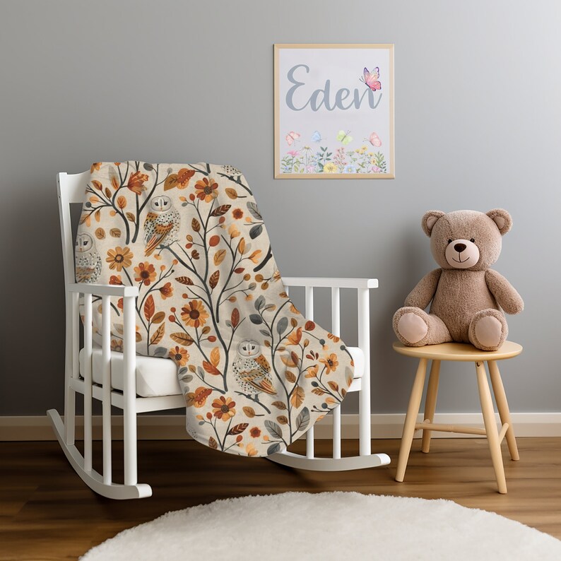 May include: A white rocking chair with a tan and orange throw blanket featuring a pattern of owls and flowers. The blanket has a cream background with brown branches and leaves. The name "Eden" is written on a framed picture above the chair.