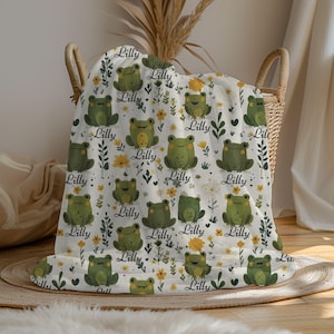 May include: A white blanket with a repeating pattern of green frogs, yellow flowers, and the name "Lilly" in cursive. The blanket is folded in a wicker basket, creating a cozy and decorative display.