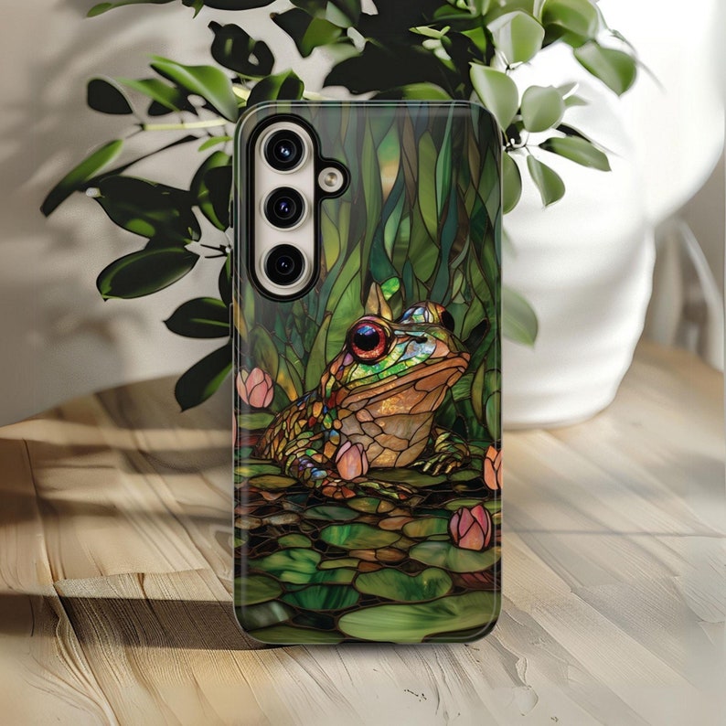 May include: A phone case with a stained glass design featuring a green frog sitting on lily pads with pink flowers. The case has a black background and a camera cutout.
