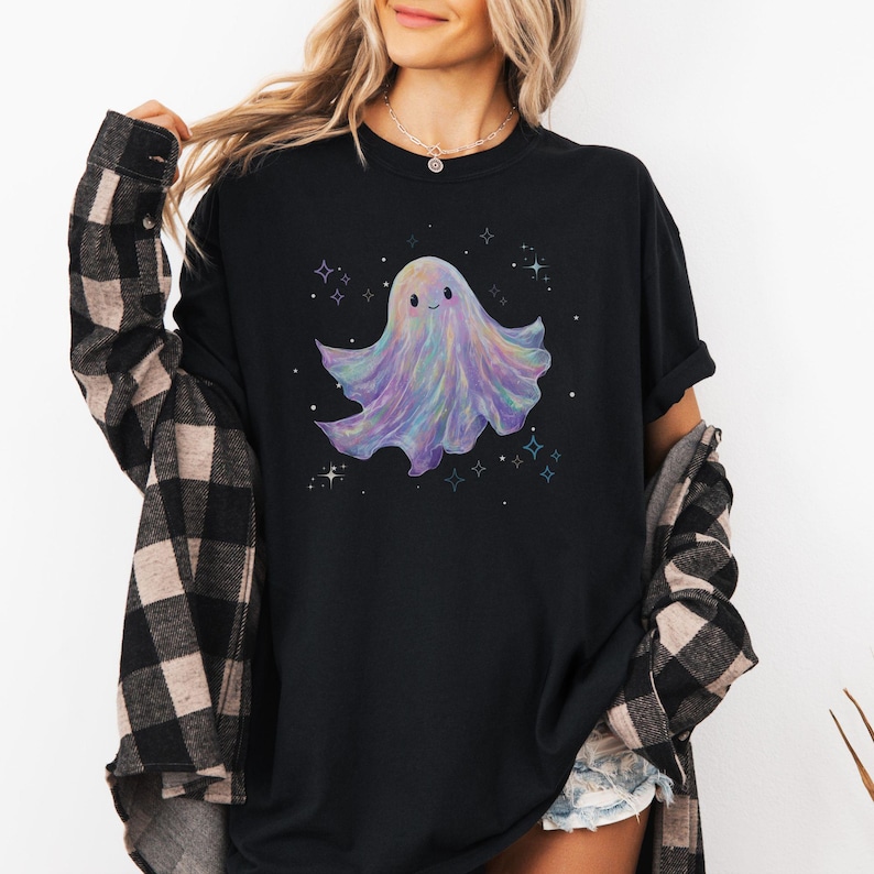 May include: Black t-shirt featuring a colorful, smiling ghost illustration. The ghost has a pastel color scheme with purple, blue, and pink hues. Small star and sparkle graphics surround the ghost, creating a whimsical design.