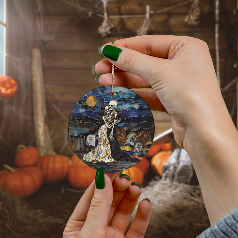 May include: Round ceramic ornament featuring a stained glass style image of two skeletons holding each other in a graveyard setting. The ornament has a gold string for hanging.