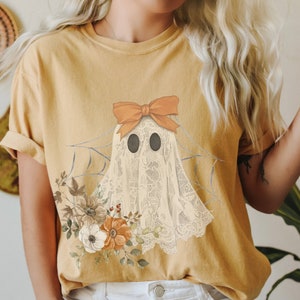 May include: A yellow t-shirt with a white ghost wearing an orange bow and surrounded by flowers. The ghost is in a spiderweb.