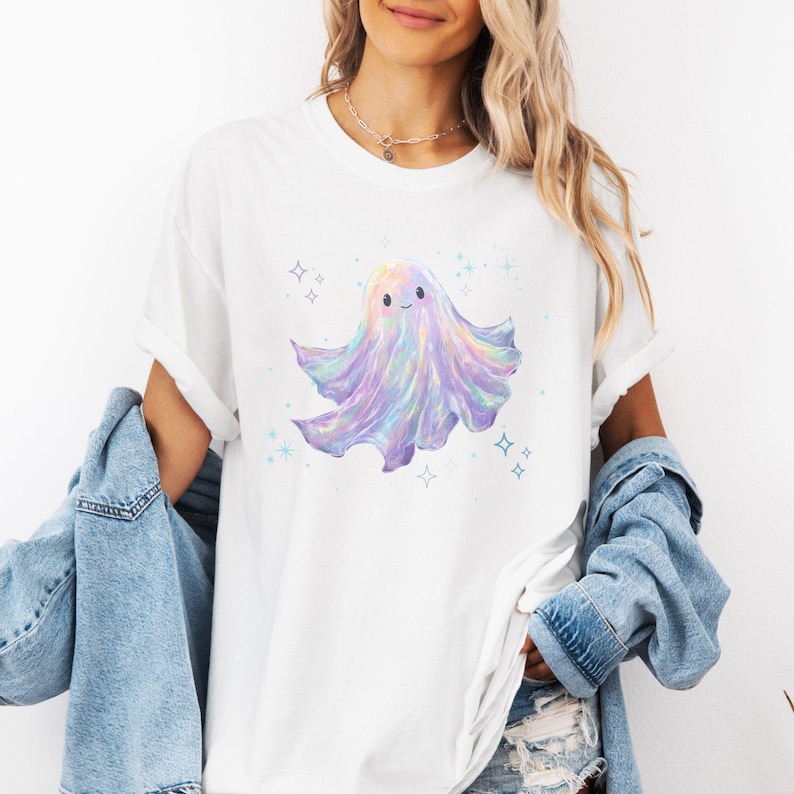 May include: White t-shirt featuring a pastel ghost graphic. The ghost has a smiling face and a flowing, iridescent body with shades of purple, blue, and yellow. Surrounding the ghost are small, sparkling star shapes.