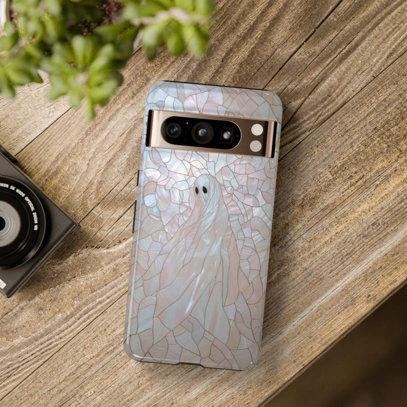 May include: A white phone case with a stained glass design featuring a ghost. The case has a gold trim around the camera lens.