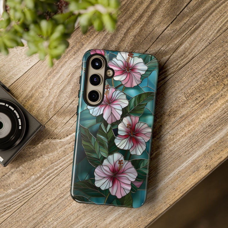 May include: A phone case with a floral design. The case features pink and white hibiscus flowers on a teal background. The phone case has a cutout for the camera lens.