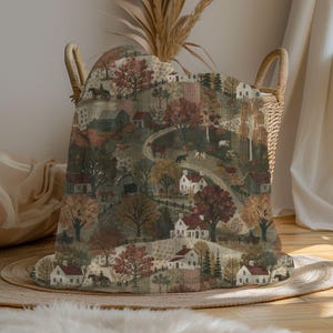 May include: A woven throw blanket featuring a repeating pastoral landscape pattern with white houses, barns, horses, and trees in shades of autumn orange, brown, and muted green. The blanket is draped over a light-colored woven wicker laundry basket.