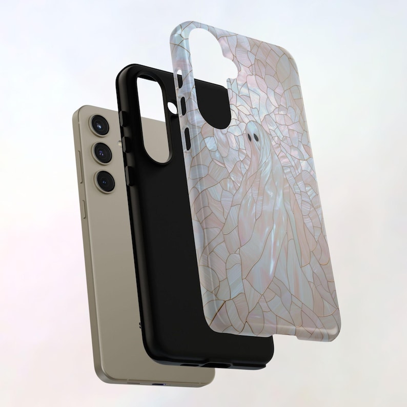 May include: A phone case with a white and pink stained glass design featuring a ghost. The case is shown with a black phone case and a gold phone.