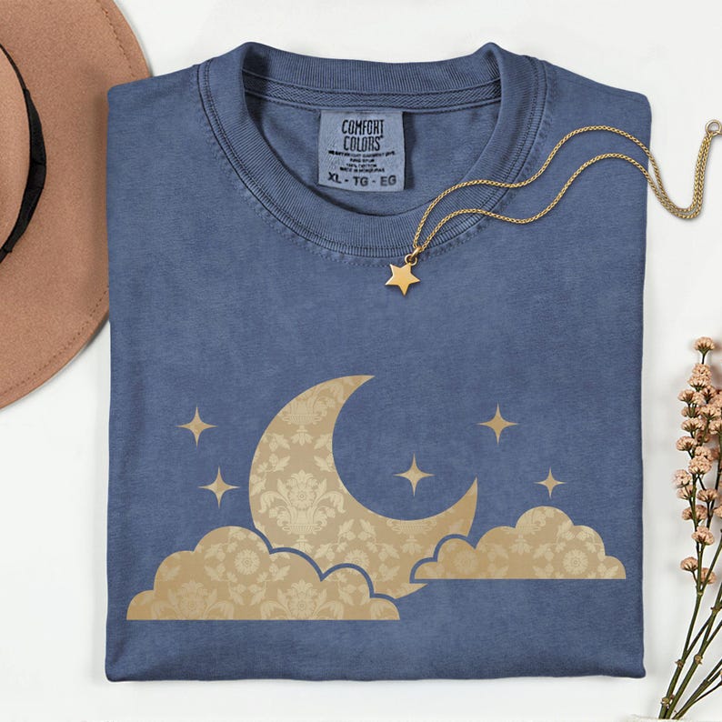 May include: A blue t-shirt with a gold crescent moon and star design. The moon and clouds have a floral pattern. A gold star necklace rests on the shirt. The shirt has a Comfort Colors tag, size XL-TG-EG.