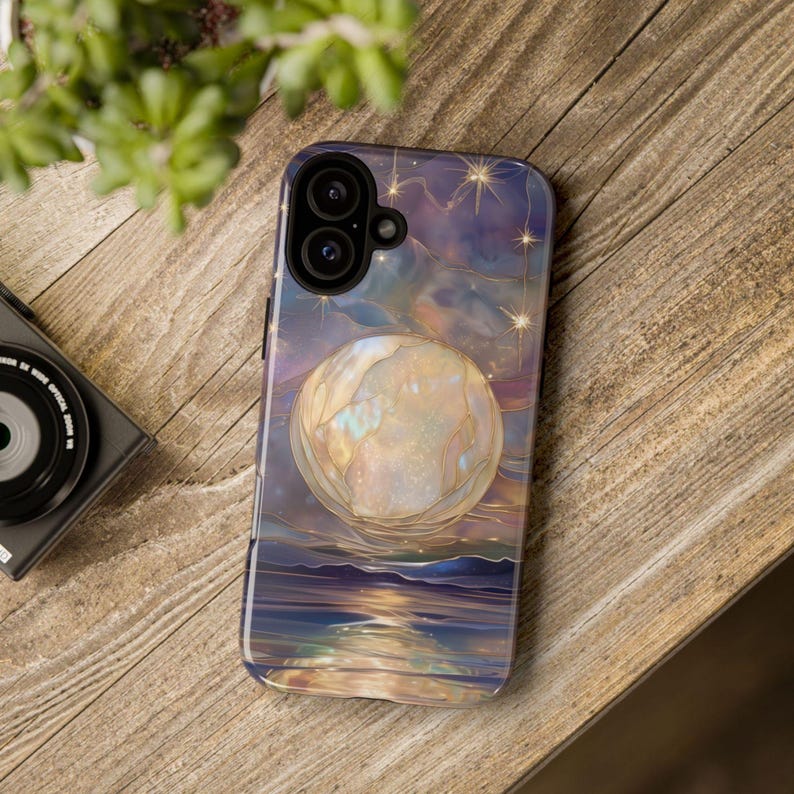 Puede incluir: Una funda de tel&eacute;fono con un dise&ntilde;o celestial, que muestra una gran luna brillante sobre el agua. La funda tiene un borde azul oscuro y un fondo de cielo estrellado en morado, azul y dorado. La funda est&aacute; sobre una superficie de madera.