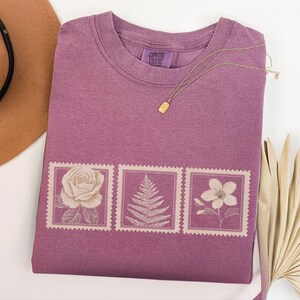 May include: A mauve Comfort Colors t-shirt with a floral stamp design. The design features a rose, fern, and flower. A gold necklace with a rectangular pendant rests on the shirt. A brown hat is visible in the upper left corner.