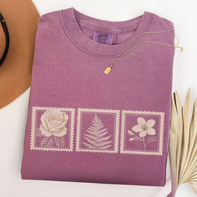 May include: A mauve Comfort Colors t-shirt with a floral stamp design. The design features a rose, fern, and flower. A gold necklace with a rectangular pendant rests on the shirt. A brown hat is visible in the upper left corner.