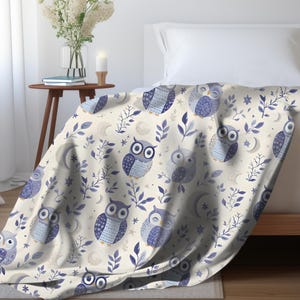 May include: A cream-colored throw blanket with a repeating pattern of blue owls and leafy branches. The owls have different expressions and are surrounded by small white stars.