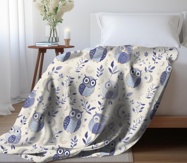 May include: A cream-colored throw blanket with a repeating pattern of blue owls and leafy branches. The owls have different expressions and are surrounded by small white stars.