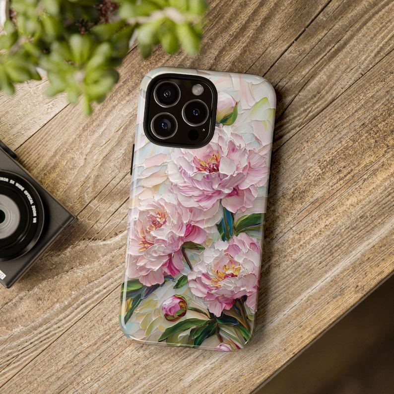 K&ouml;nnte beinhalten: Eine Handyh&uuml;lle mit einem Blumendesign mit rosa Pfingstrosen auf wei&szlig;em Hintergrund. Die H&uuml;lle hat einen Kameraausschnitt mit drei Objektiven.