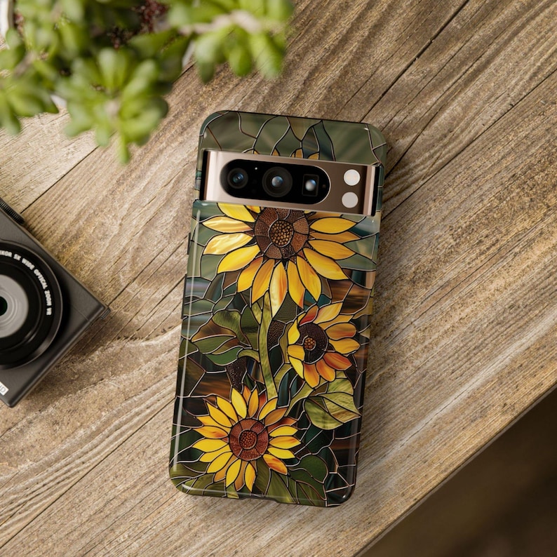 May include: A phone case with a stained glass design featuring three yellow sunflowers with brown centers. The case has a camera cutout with a gold trim.