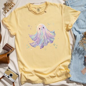 May include: A yellow t-shirt featuring a cute, colorful ghost illustration. The ghost has a smiling face and a flowing, iridescent body with shades of purple, blue, and pink. The shirt is styled with a gold necklace and denim jeans.