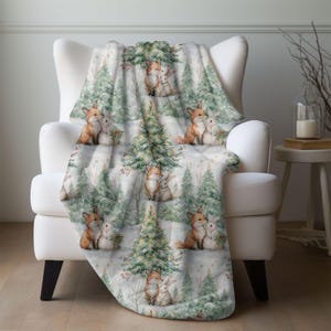 May include: A soft, white blanket draped over a white armchair. The blanket features a repeating pattern of foxes and rabbits nestled together, surrounded by green Christmas trees. The blanket is a cozy home decor item.