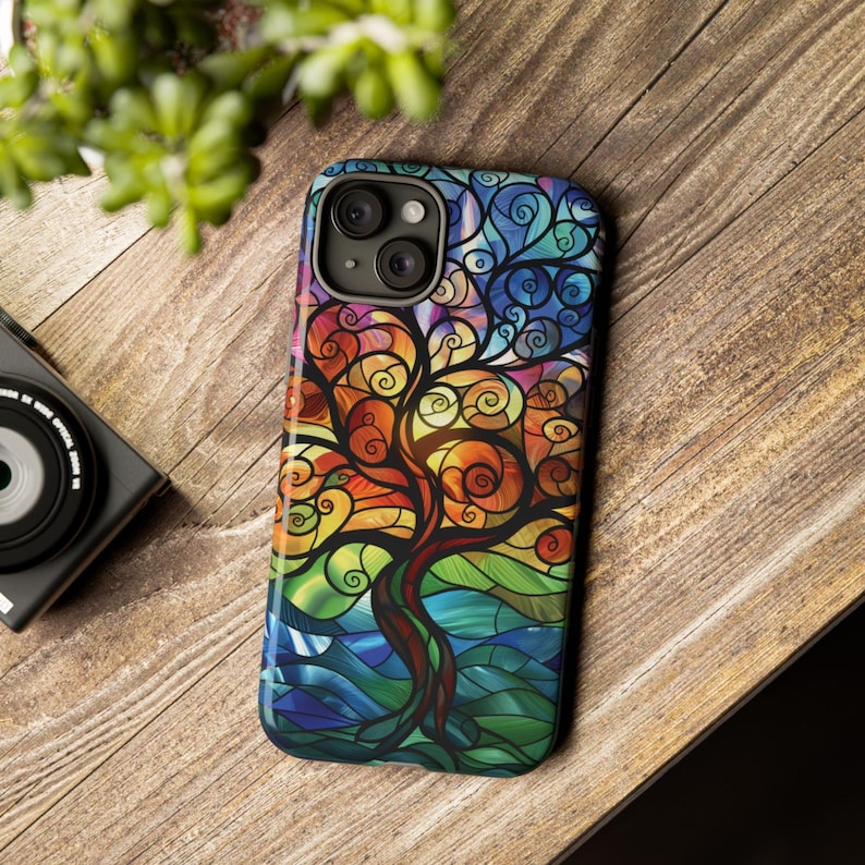 May include: A colorful phone case with a stained glass design of a tree with swirling branches. The case is made of durable plastic and features a raised camera bump for protection.