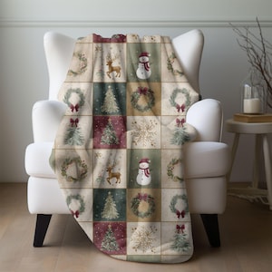 May include: A Christmas-themed throw blanket draped over a white armchair. The blanket features a patchwork design with squares of wreaths, reindeer, snowmen, Christmas trees, and snowflakes in shades of red, green, and beige.