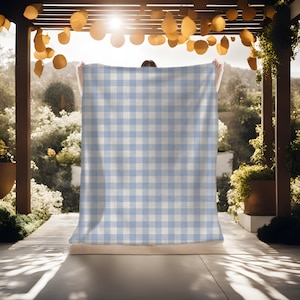 May include: A light blue and white gingham check picnic blanket. The blanket is being held up by a person in a sunny outdoor setting.