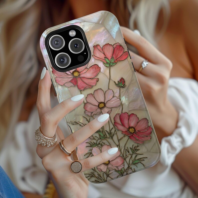 May include: A decorative phone case featuring a floral design with pink cosmos flowers and green stems. The case has a pearlescent background and a protective bumper around the camera.