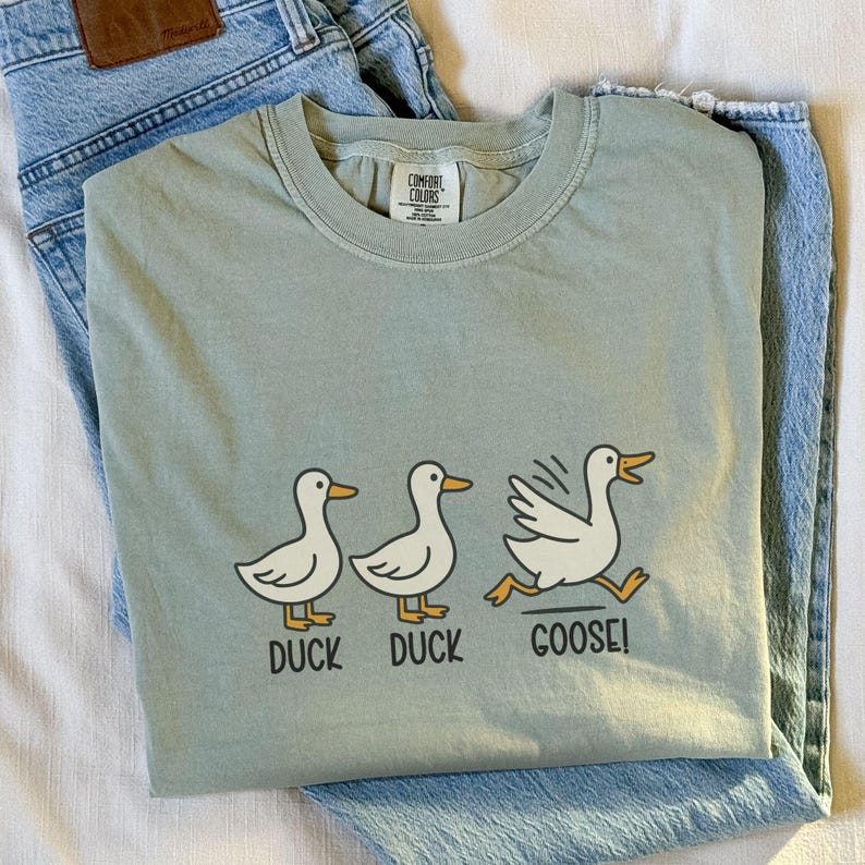 May include: Sage green t-shirt with a graphic of three cartoon ducks. The first two ducks are standing, and the third is running with the text "DUCK DUCK GOOSE!" below them. The shirt is laid on top of blue jeans.