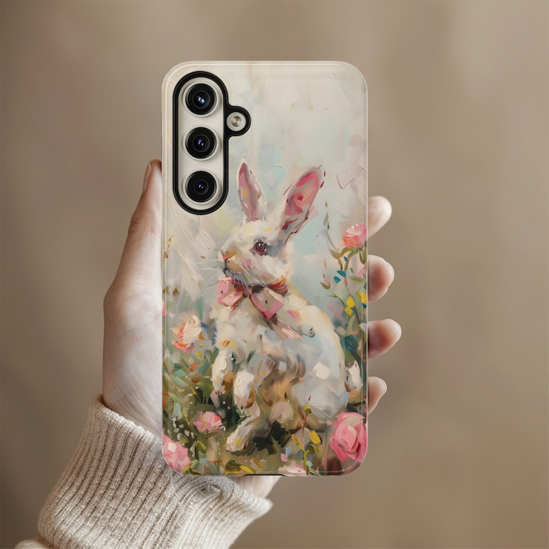 K&ouml;nnte beinhalten: Ein wei&szlig;es Kaninchen mit einer rosa Schleife, das auf einer Handyh&uuml;lle mit floralem Hintergrund gemalt ist. Die Handyh&uuml;lle hat ein dreifaches Kameraobjektiv.