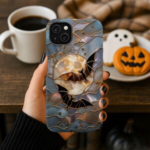Pu&ograve; includere: Una custodia per telefono con un design in stile vetrata che raffigura una luna piena, due pipistrelli neri e piccole stelle. Lo sfondo ha una finitura madreperlacea e iridescente nei toni del blu, grigio e bianco, con sottili linee dorate.
