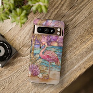 Puede incluir: Una funda de tel&eacute;fono con un dise&ntilde;o de flamenco. La funda tiene una colorida ilustraci&oacute;n de un flamenco rosa en una playa con un atardecer y un fondo de oc&eacute;ano. La funda est&aacute; dise&ntilde;ada para un tel&eacute;fono con c&aacute;mara y flash.