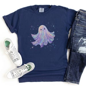 May include: Navy blue t-shirt featuring a whimsical, pastel-colored ghost illustration with sparkling stars. The ghost has a friendly expression. The shirt is paired with blue jeans and gray sneakers, creating a casual, everyday look.