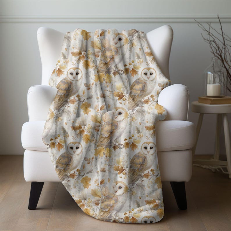 May include: A cream-coloured throw blanket with a repeating pattern of owls, branches, and autumn leaves in shades of brown, tan, and gold. The blanket is draped over a white armchair. The blanket is made of a plush material.