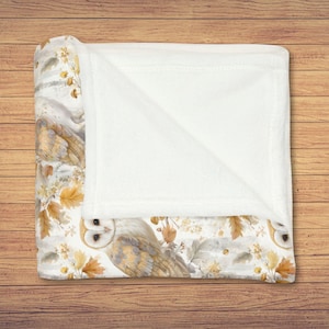 May include: A folded blanket with a white and patterned design. The blanket features a watercolor design of owls, autumn leaves, and floral elements in shades of brown, beige, and yellow. The blanket is folded over, revealing a soft, white, plush interior.