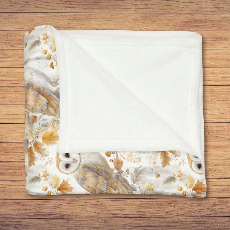May include: A folded blanket with a white and patterned design. The blanket features a watercolor design of owls, autumn leaves, and floral elements in shades of brown, beige, and yellow. The blanket is folded over, revealing a soft, white, plush interior.