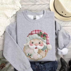 May include: A gray sweatshirt featuring a Santa Claus applique. The Santa design includes a red and green plaid hat, a white beard, and a rosy-cheeked face. The sweatshirt is paired with blue jeans, a white hat, and black boots.