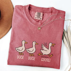 May include: A red-pink Comfort Colors t-shirt with a crew neck and the words "DUCK DUCK GOOSE!" printed below three cartoon ducks. A silver necklace with a small pendant is draped across the shirt.