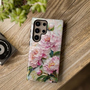 K&ouml;nnte beinhalten: Eine Handyh&uuml;lle mit einem Blumendesign mit rosa Pfingstrosen und gr&uuml;nen Bl&auml;ttern. Die H&uuml;lle hat einen Kameraausschnitt mit schwarzem Rand.