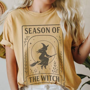 May include: Mustard-colored t-shirt featuring a black graphic of a witch on a broomstick inside a rectangular frame. The text "SEASON OF THE WITCH" is printed above and below the witch illustration. The shirt has short sleeves.