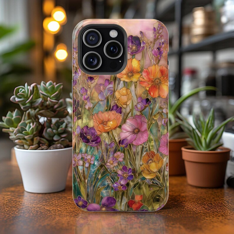 Op de afbeelding: Een telefoonhoesje met een levendig bloemmotief met oranje, paarse en roze bloemen. De hoes heeft een glanzende afwerking en een beschermende bumper. Het ontwerp lijkt op een glas-in-loodraam met ingewikkelde details en een kleurrijk palet.