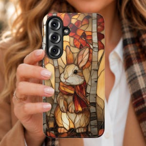 May include: A phone case featuring a stained glass style illustration of a rabbit wearing a scarf. The design incorporates warm autumn colors including orange, brown, and yellow, with a forest background of trees. The case has a black camera cutout.