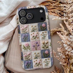 May include: A floral phone case featuring a patchwork design with embroidered flowers in pink, purple, and white. The case has a black camera module and a soft, neutral background.