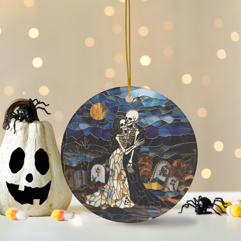 May include: A round ceramic ornament with a stained glass design of two skeletons embracing in a graveyard setting. The skeletons are dressed in a white gown and a black robe. The background is a dark blue sky with a full moon.