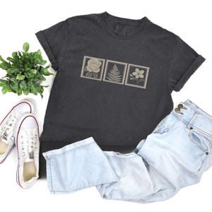 May include: Dark gray t-shirt with a floral stamp design featuring a rose, fern, and flower. The shirt is paired with light blue jeans and white sneakers with red accents. A small green plant is also in the image.