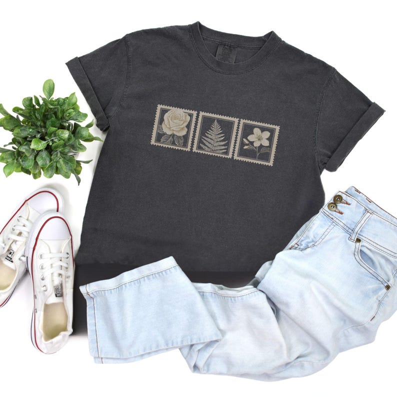 May include: Dark gray t-shirt with a floral stamp design featuring a rose, fern, and flower. The shirt is paired with light blue jeans and white sneakers with red accents. A small green plant is also in the image.