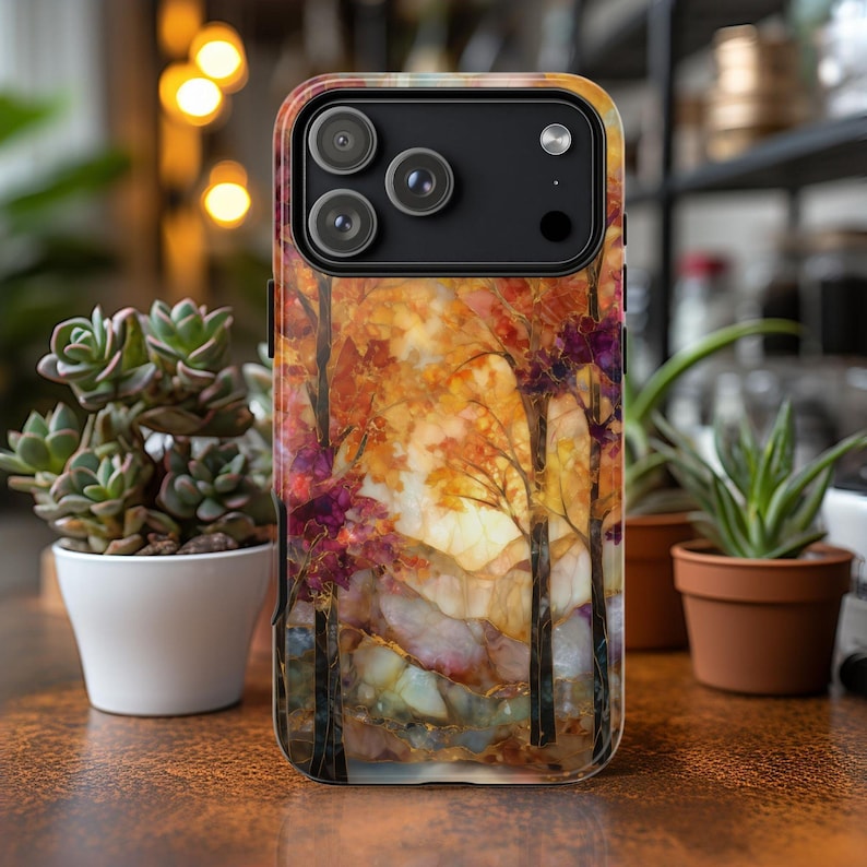 May include: A phone case featuring an autumn landscape design. The case showcases a vibrant scene with trees in shades of orange, red, and purple, set against a warm, golden background. The design includes a mountain range and a touch of gold.